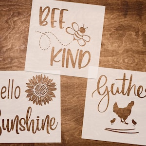 May include: Three white stencils with black lettering and images. The first stencil says "Bee Kind" with a bee image. The second stencil says "Hello Sunshine" with a sunflower image. The third stencil says "Gather" with a hen and chicks image.
