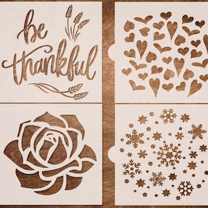 May include: Four white stencil designs on a wood background. The designs are "be thankful" with wheat stalks, a cluster of hearts, a rose, and a snowflake pattern.