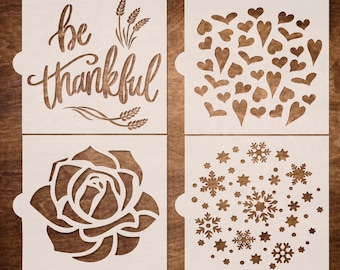 May include: Four white stencil designs on a wood background. The designs are "be thankful" with wheat stalks, a cluster of hearts, a rose, and a snowflake pattern.