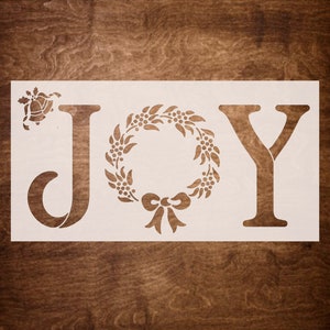 May include: White stencil with the word "JOY" in a decorative font. The "O" is replaced with a wreath with a bow. A small bell with a holly sprig is to the left of the "J".