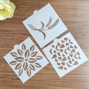 May include: Three white plastic stencils with different designs. The first stencil has a wheat stalk design. The second stencil has a floral design. The third stencil has a design of many small hearts.