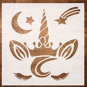 May include: A white unicorn stencil with a crescent moon, star, and shooting star design. The stencil features a unicorn horn, crown, and closed eyelashes. The stencil is made of a light-colored material and is set against a brown wooden background.