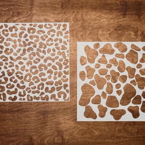 May include: Two white stencils on a brown wooden background. The stencil on the left has a leopard print design. The stencil on the right has a pattern of irregular circles.