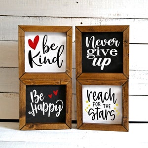 May include: Four wooden framed signs with inspirational messages. The signs are white and black with red and gold lettering. The messages are "Be Kind", "Never Give Up", "Be Happy", and "Reach for the Stars".