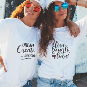 May include: Two women wearing white t-shirts with different inspirational messages. The woman on the left has a t-shirt that says "Dream Create Inspire". The woman on the right has a t-shirt that says "Live Laugh Love" with small red hearts around the text.