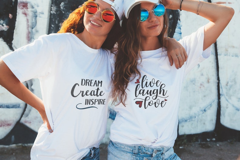 May include: Two women wearing white t-shirts with different inspirational messages. The woman on the left has a t-shirt that says "Dream Create Inspire". The woman on the right has a t-shirt that says "Live Laugh Love" with small red hearts around the text.