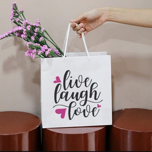 May include: White paper gift bag with pink hearts and the text "live laugh love" in black script font.