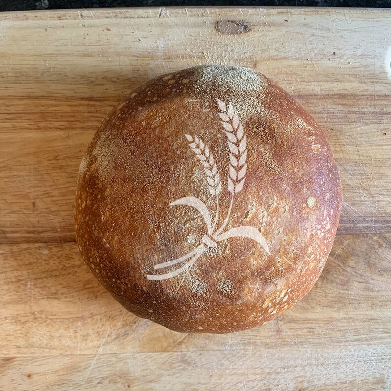 May include: A round loaf of sourdough bread with a wheat design on the top.