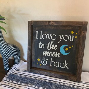 May include: A wooden sign with a dark brown frame and a black background. The sign says "I love you to the moon & back" in white lettering with a blue crescent moon and yellow stars.