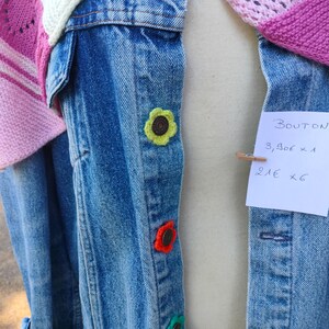Peut inclure: Une veste en jean bleu ornée de quatre couvre-boutons en forme de fleurs crochetées aux couleurs rose, jaune, orange et vert. Une étiquette en papier blanc est fixée par une pince en bois avec le texte BOUTON 3,90€ x 1 21€ x 6.