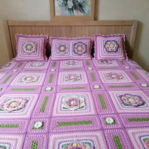 May include: A crocheted bedspread with a floral pattern in shades of pink, white, and green. The bedspread is on a bed with three matching crocheted pillows.