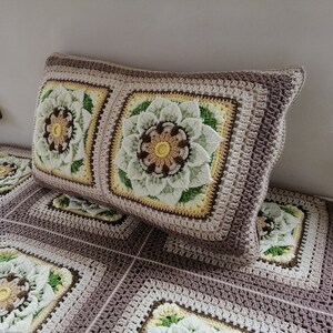 May include: A crocheted pillow with a floral design in shades of brown, green, and white. The pillow is made of a soft, textured yarn and has a square shape.