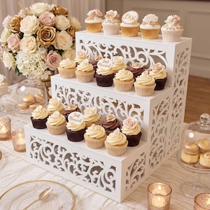 Op de afbeelding: Een witte cupcake stand met drie niveaus en decoratieve uitsparingen toont cupcakes met bloemen en "Just Married" toppers. Een boeket bloemen in goud, roze en wit staat achter de stand. Andere desserts en kaarsen zijn ook zichtbaar.