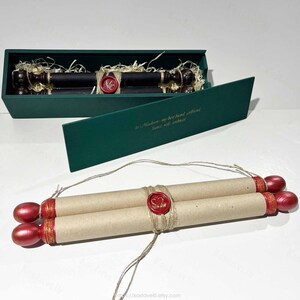 Op de afbeelding: Een beige papieren rol met rood geschilderde houten uiteinden en een rood waszegel, vastgebonden met touw. Het ligt voor een donkergroene rechthoekige doos met een soortgelijke rol erin, met gouden tekst op het deksel.
