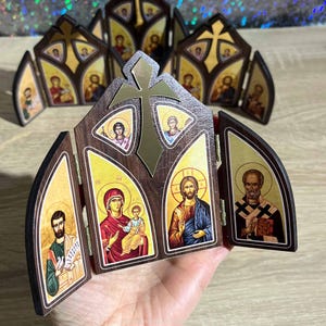 May include: Wooden religious icon featuring images of saints and religious figures. The icon has a dark brown wooden frame with a gold cross and arched panels. The images are colorful and detailed, with gold backgrounds.