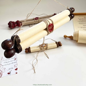 May include: A set of rolled parchment scrolls featuring dark wooden finials at the ends, secured with twine and red wax seals. The scrolls are suspended by thin string, with additional scrolls and a printed card with text visible in the background.