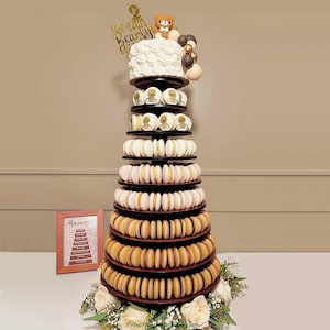May include: A multi-tiered macaron tower in shades of brown, white, and pink, topped with a white cake, a teddy bear, and a gold topper reading "we can bearly wait". The base is decorated with white roses. The tower is a visually appealing dessert.