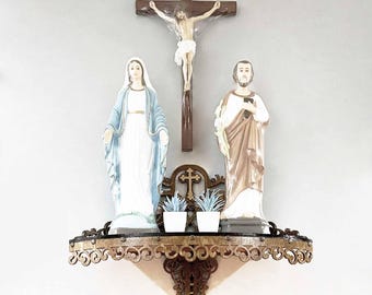 May include: A wall-mounted dark brown wooden shelf with ornate scrollwork edges holds two religious statues and two small white pots with artificial plants. A crucifix is mounted above the shelf between the statues of Mary and Joseph.