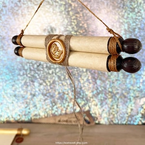 May include: Two rolled parchment scrolls, secured with twine, and finished with dark wooden finials and a gold wax seal. The scrolls are suspended by a natural fibre cord, creating a vintage aesthetic. The seal has a detailed embossed design.