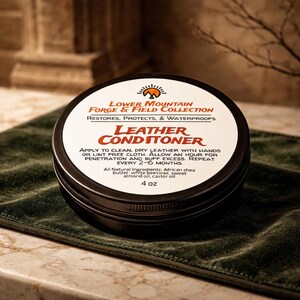 May include: A round, black metal tin of "Leather Conditioner" from the Lower Mountain Forge & Field Collection. The label is white with red and black text. The tin is sitting on a dark green cloth. The product contains 4 oz.