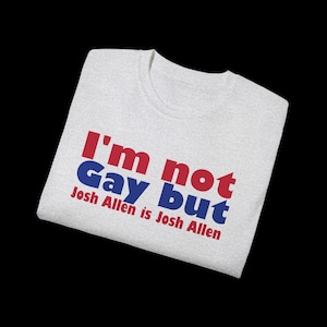 May include: A light gray t-shirt with the text "I'm not Gay but Josh Allen is Josh Allen" printed in red and blue. The shirt is folded and displayed against a black background.