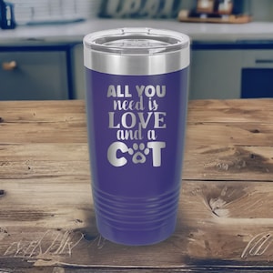 May include: Purple stainless steel tumbler with a silver lid. The tumbler has the text "All you need is love and a cat" with a paw print replacing the "o" in cat.