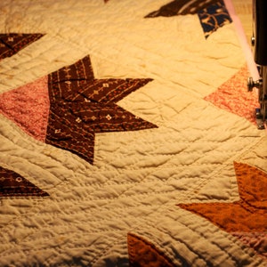 May include: A close-up of a quilt being sewn on a sewing machine. The quilt is made of various fabrics in shades of brown, pink, and orange. The quilt is being sewn with a pink binding.