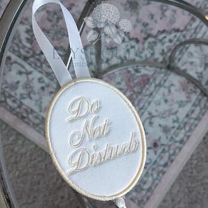 May include: An oval-shaped white fabric door hanger featuring the embroidered text "Do Not Disturb" in a gold script font. The hanger includes a gold-stitched border, a white ribbon loop for hanging, and a white tassel attached to the bottom edge.