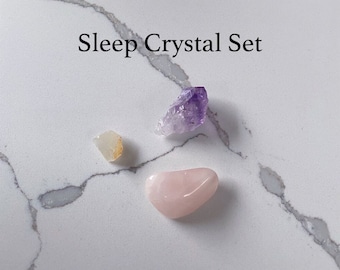 Puede incluir: Un conjunto de tres cristales para dormir, que incluye una amatista morada, un cuarzo rosa y un citrino claro. Los cristales están dispuestos sobre una superficie de mármol blanco. Juego de cristales para dormir