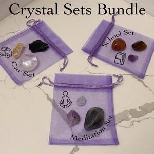 May include: Three purple organza bags with different crystal sets. The first bag says "Car Set" and contains a yellow crystal, a black crystal, and a white crystal. The second bag says "School Set" and contains a red crystal, a brown crystal, and a purple crystal. The third bag says "Meditation Set" and contains a green crystal, a purple crystal, and a clear crystal.