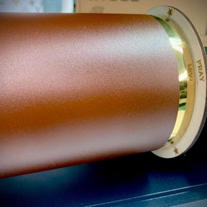 Puede incluir: Primer plano de un rodillo marr&oacute;n con una banda dorada y una tapa de madera. La tapa tiene la palabra "PRAY" escrita en ella.