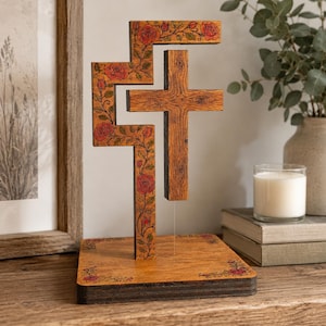 Puede incluir: Una escultura de cruz de madera con una sección transversal flotante suspendida sobre una base. La estructura está decorada con un patrón detallado de rosas rojas y enredaderas verdes pirograbadas. La base es un cuadrado de madera plano con arte floral a juego.