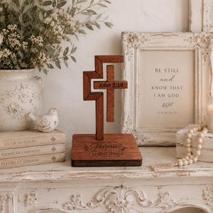 Puede incluir: Una escultura de cruz de madera marrón oscuro montada sobre una base cuadrada. La cruz presenta el texto grabado John 3:16 en la barra horizontal y el nombre Jennie con la frase He sent Jesus grabada en la base, creando un objeto religioso.
