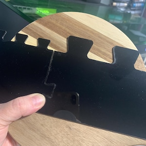 May include: A wooden coaster with a wavy edge and a black acrylic insert. The insert has a series of interlocking pieces.