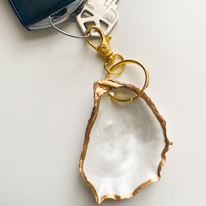 May include: A white oyster shell keychain with a gold-painted edge and a gold-colored clasp. The keychain is attached to a car key and a house key.