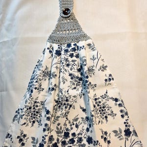 May include: A hanging kitchen towel with a gray crocheted top and a button closure. The towel features a white background with a floral pattern in shades of blue and gray. The towel is designed to hang from a hook.