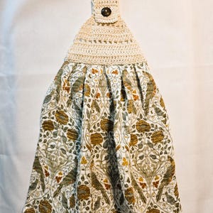 May include: Hanging kitchen towel with a cream-colored crocheted top and a button closure. The towel portion features a floral pattern in shades of green, brown, and gold on a white background. A decorative and functional kitchen accessory.