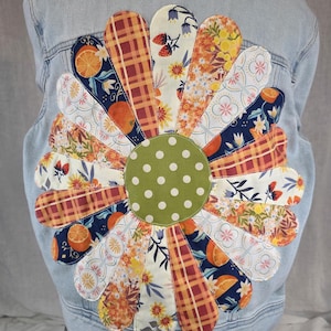 May include: A light blue denim jacket featuring a large, circular flower appliqué on the back. The flower has a central olive green circle with white polka dots, surrounded by numerous petals made from various fabrics with orange, plaid, and floral prints.