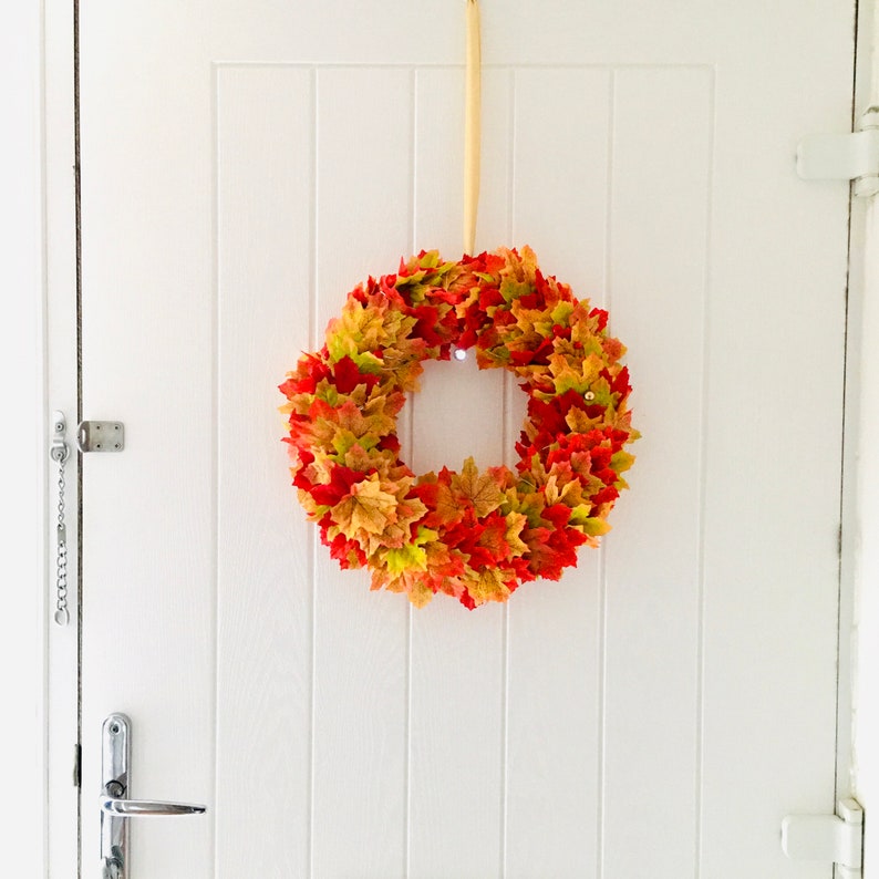 May include: A fall-themed wreath made of artificial leaves in shades of orange, red, and yellow. The wreath is hanging on a white door.