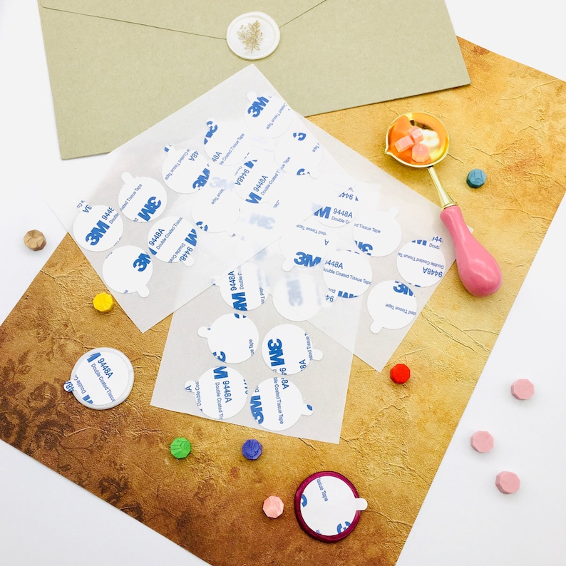 May include: A close-up of a sheet of white paper with multiple round, white adhesive stickers. The stickers are labeled with the text "3M 9448A Double Coated Tissue Tape". The stickers are arranged in a random pattern on the paper. There are also several small, colorful wax seals scattered around the paper.