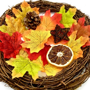 May include: A fall-themed wreath with a brown vine base decorated with orange, yellow, and red maple leaves, a pine cone, a dried orange slice, and a woven star.