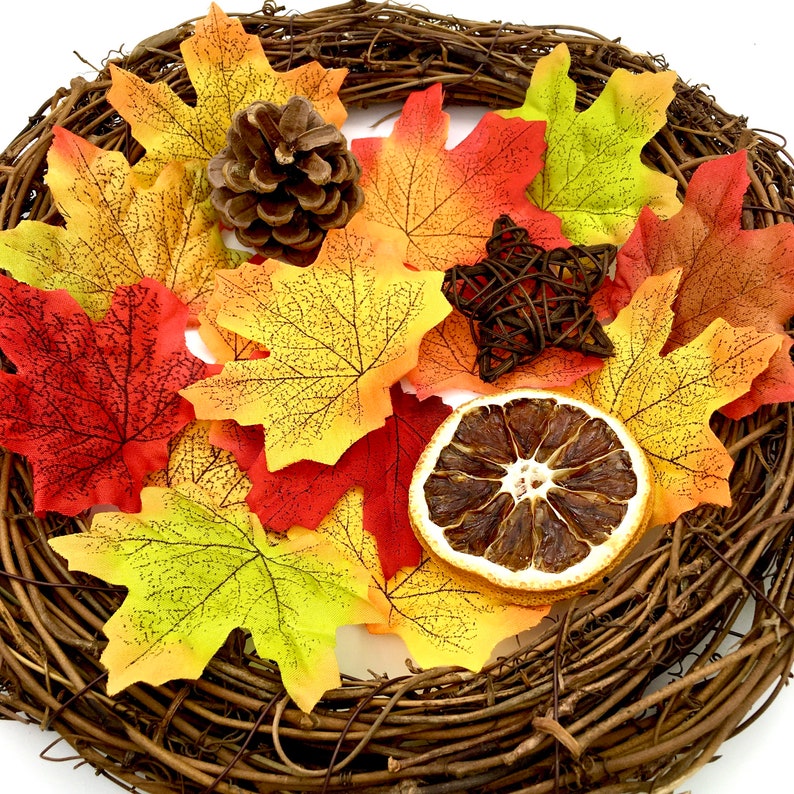 May include: A fall-themed wreath with a brown vine base decorated with orange, yellow, and red maple leaves, a pine cone, a dried orange slice, and a woven star.