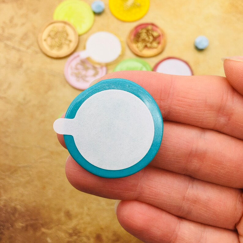 May include: A teal round sticker with a white circle in the center. The sticker has a small tab on the side.