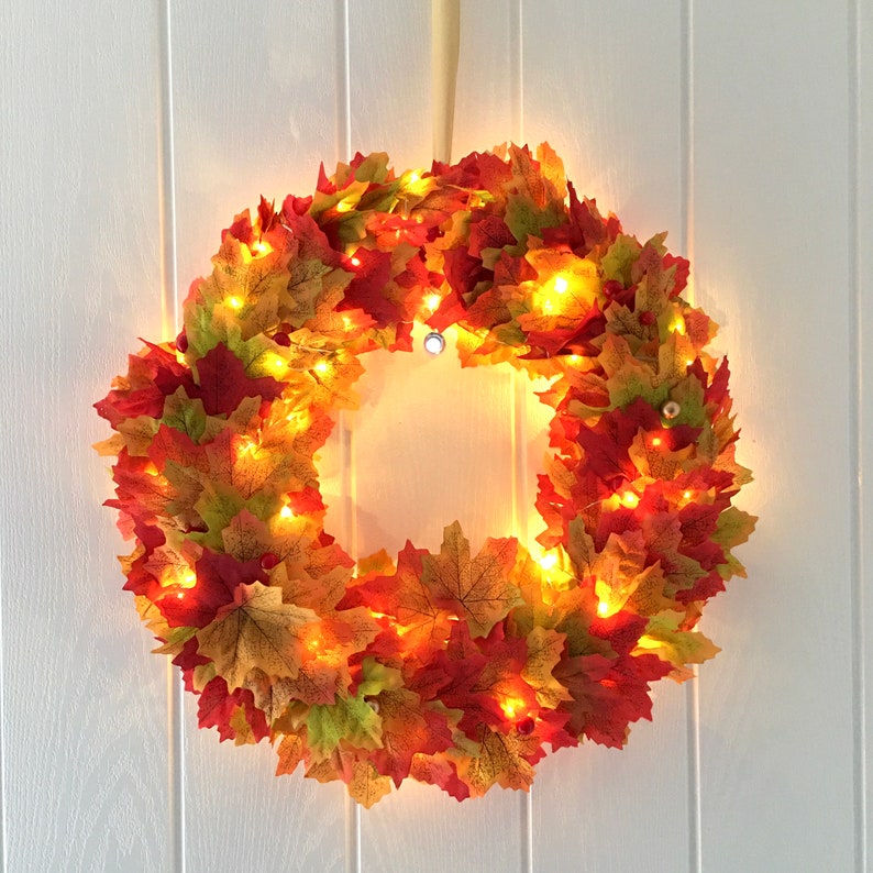 May include: A fall-themed wreath made of artificial leaves in shades of red, orange, and yellow. The wreath is decorated with warm white fairy lights.