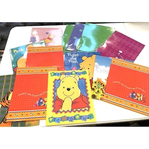 May include: A collection of Winnie the Pooh themed stationery items. The items include notebooks and cards with illustrations of Pooh, Piglet, and Tigger. The notebooks have orange and red backgrounds with decorative borders. The cards feature various scenes and characters.
