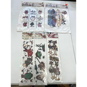 May include: Five packages of iron-on fabric transfers featuring various designs. The top two show apple patterns and a person in a hat. The bottom three display angels, fishing gear with a fish, and birds with sunflowers. Each package is sealed in clear plastic.