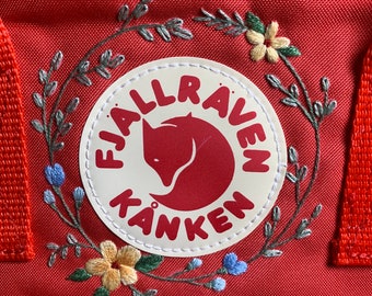 Op de afbeelding: Rode Fjällräven Kånken rugzak met een witte geborduurde patch met een vos en de merknaam. De patch is omringd door een krans van groene bladeren en gele en blauwe bloemen.