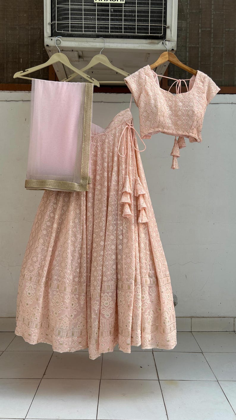 May include: A light pink, embroidered lehenga set, including a skirt, blouse, and dupatta. The skirt and blouse feature intricate patterns. The dupatta has a gold border. The set is hanging on wooden hangers.