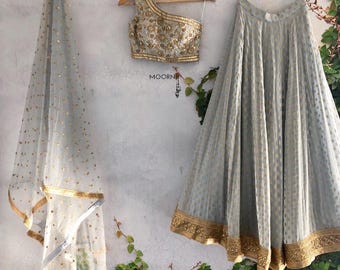 Puede incluir: Un top corto de un solo hombro gris y dorado bordado con una falda gris a juego y una dupatta gris transparente con detalles dorados. El atuendo está colgado en perchas contra una pared blanca con enredaderas verdes.
