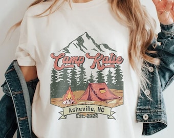 May include: White t-shirt with a vintage-style graphic of a mountain range, a tent, a campfire, and the text "Camp Katie Asheville, NC Est. 2024".